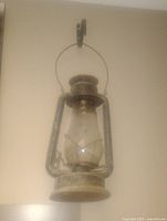 Image shows an antique metal lantern with a glass globe, hanging on a wall. The lantern has a metal handle and a wire frame around the glass.