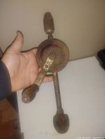 Front view of the antique hand drill tool showing circular gear, wooden top handle, and wooden crank handle, held in hand for scale.