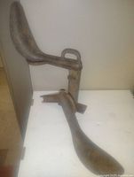 Photo showing large metal shoemaker's last with curved foot and handle, resting on surface with smaller tool beside it.
