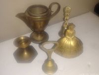 Photo of five brass items on white surface including a small ornate pitcher, figurine bell, goblet, candle holder, and bottle opener.
