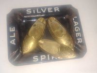 Three brass mini shoe-shaped ashtrays arranged inside a black metal beer tray with white text that reads 'ALE SILVER LAGER SPI'. Shoes have detailed design and polished brass appearance.