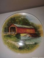 Round vintage plate with painted image of red covered bridge over creek with greenery and rowboat