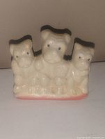 Photo showing ceramic figurine of three dogs in cream with black and gray painted details on a pink base.