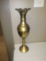 Image of a large brass vase with a flared scalloped rim and bulbous midsection, placed against a light wall.