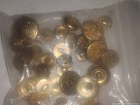 Plastic bag containing multiple brass-colored military buttons with embossed designs and symbols, various sizes.