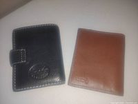 Photo showing two leather wallets: one black with a snap button clasp and white stitching, the other brown bi-fold wallet. Both have embossed logos on the front and appear lightly used.