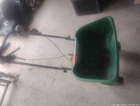 Green seed spreader with large hopper, black handle, and wheels indoors on concrete floor.