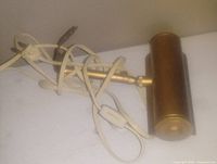 Photo showing a vintage brass cylindrical painting lamp with attached white power cord and inline switch, positioned on a white surface.