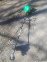 Full length view of green gas powered weedeater lying on dirt ground, showing 65 inch shaft with handle loop and cutting head with guard.