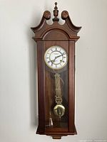 Front view of the wooden Waltham regulator wall clock with glass door, carved finial top, and visible brass pendulum inside.