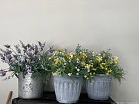 Three artificial flower arrangements; two in gray plastic pots and one in silver aluminum pot, showing full set and details of flowers and foliage.