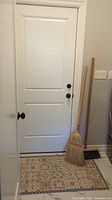 Two brooms and a vinyl floor mat placed leaning against a white door on patterned tile floor.