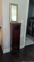 Tall narrow dark wood veneer cabinet with glass window upper door and bottom drawer against wall under a rectangular mirror with gold crackle frame.