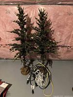 Two rustic artificial Christmas trees with lights, burlap-wrapped bases, and electrical cords on gray floor against pink insulation wall.