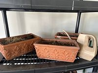 View of three terracotta clay rectangular planters with decorative leaf pattern set on a black shelf, along with a beige plastic watering can.