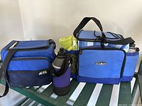 Photo showing two blue Arctic Zone coolers, two water bottles with neoprene sleeves, and a green towel on a glass surface against white wall.