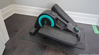 Side angle of the full seated elliptical machine on a black mat near a baseboard wall, showing the foot pedals and body of the machine.