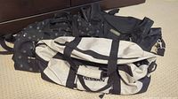 Three duffle style bags placed on carpet floor: One white and black Nissan branded bag showing marks from use, plus two black duffle bags with small pattern print and multiple zippered pockets.