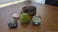 Four items on wood surface: three paperweights (two glass with floral designs, one acrylic with Milk Weed seed) plus purple amethyst geode in rear
