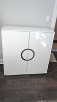 Front view of high gloss white cabinet showing 4 closed doors with round mirrored handles forming a circle in the center.