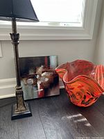 Wide view showing the lamp, abstract art canvas, and large orange-red glass planter positioned together on the floor by a window.