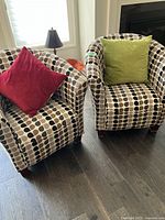 Two upholstered tub chairs with geometric patterned fabric, one with a red pillow and the other with a green pillow, positioned on dark hardwood floor.