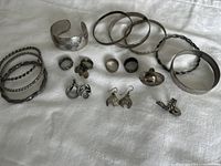 Wide view of assorted silver tone jewelry laid on white fabric, showing bracelets, rings, and earrings.