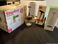 Starfrit Spiralizer in box with a hand chopper glass with brown lid, a vacu vin pineapple slicer box, and a Prepara herb keeper box