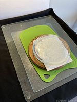 Overview of four kitchen items stacked: clear textured glass cutting board, green plastic cutting board, round wooden cutting board, and round translucent glass/ceramic hot plate
