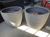 Two large patio planters side by side on a concrete floor with some furniture in background, showing rough textured, heavy build.