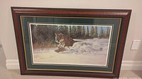 Framed print of cougar running through snow with forest background, showing entire piece including frame and matting.