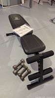 Full view of Marcy utility bench in black with manual on seat and two metal hand weights (8lb and 5lb) on the floor.