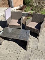 Two woven resin armchairs with beige cushions and the rectangular woven resin table with glass top placed on stone patio outdoors
