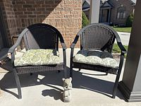 Two black patio chairs with dark woven seats and backs, each with green and white floral cushions, shown outside on concrete against brick home wall.