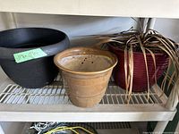 Three different planters on white metal shelf: two black plastic planters, one ceramic planter, one large red plastic planter with dry plant inside