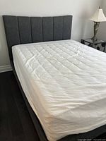 Full view of the queen bed with upholstered headboard, white covered mattress and base on dark flooring.