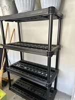 Full view of the black plastic shelf unit with four tiers, assembled and standing upright against a wall. Various items are partially visible around it including brooms and storage containers.