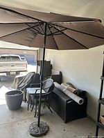 Full view of the open dark colored sun umbrella with metal ribs and black pole standing on a cement base inside a garage or covered area.