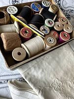 Box of assorted vintage thread spools in various colors including black, white, red, and purple, both wooden and plastic spools.