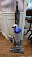 Full view of Dyson upright vacuum cleaner with included accessories (spot cleaner spray and maintenance powder) placed on wooden floor against wall.
