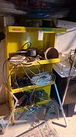 Yellow shelving unit with assorted electrical wires, cords, adapters, and small tools in basement setting.