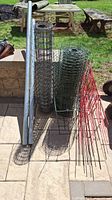 Photo showing all items: two rolls of wire fencing (one galvanized, one green coated), multiple red metal plant stands or garden stakes, and two cylindrical metal poles lying next to fencing on paved surface outdoors.