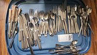 Top-down view of assorted silver-tone flatware including forks, knives, spoons arranged on a blue tray, with visible metal handles and wooden handled knives.