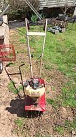 Front view of the red Mantis tiller showing handlebars, motor, and tines against a grassy outdoor background.