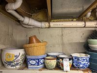 Seven assorted planters and flowerpots in various sizes, shown on shelving against concrete wall in basement.