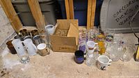 Wide view of assortment of glasses and mugs on basement floor including milk glass and branded collectible mugs