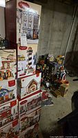 Stacked boxes of Saint Nicholas Square village buildings including Daisy's Florals & Gifts, lighthouse, and other village structures in a basement setting.