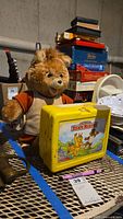 Teddy Ruxpin plush, lunchbox and thermos placed on a table with various other items in background.