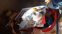 Plastic basket with assorted metal and plastic tools including grease guns, sanding tools, and electrical accessories in a red bucket.