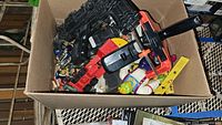 Multiple black and red plastic toy guns and various small toys in cardboard box
